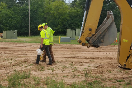 Two students wearing high-visibility jackets and hard hats walk near an excavator on a worksite.