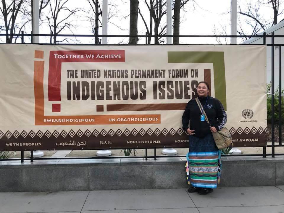 Kemewan Waupekenay standing in front of a banner for the United Nations Permanent Forum on Indigenous Issues.