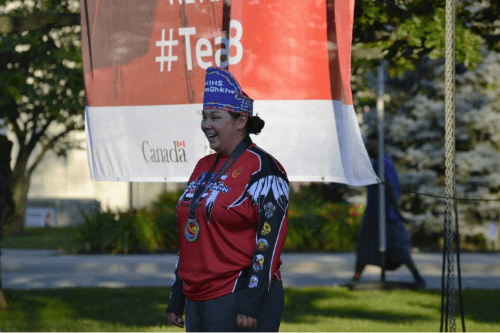 A woman wearing red archery gear and a medal around her neck smiles while stand under a banner outside.