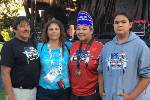   Four individuals standing together outdoors, smiling at the camera. They are wearing various shirts with logos, including "Team Wisconsin." Two attendees have lanyards around their necks. Trees and a stage setup are visible in the background.