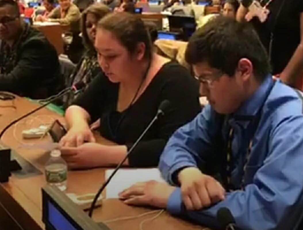 Kemewan Waupekenay speaking at a microphone in a United Nations conference room, with attendees listening in the background.