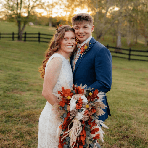 Two individuals in wedding attire smiling outdoors, one holding a bouquet of vibrant flowers. A fenced meadow at sunset forms the background.