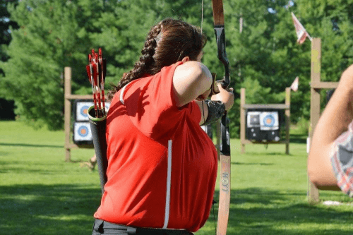 An archer in a red shirt aiming a bow at a target in a sunny outdoor archery range.