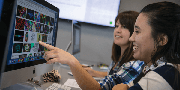 A student points to a computer screen while giving a demo to another student