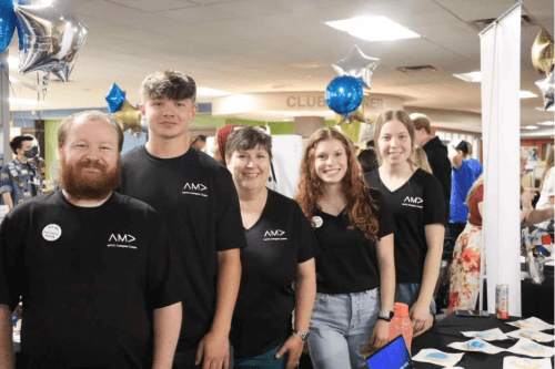 Five individuals wearing AMA t-shirts standing at a promotional booth with balloons in the background.